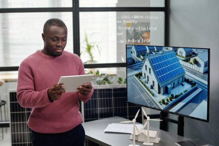Téléchargez les photos : Jeune homme d'affaires sérieux regardant écran tablette contre son lieu de travail avec tableau interactif et des modèles de moulins à vent et de la maison - en image libre de droit