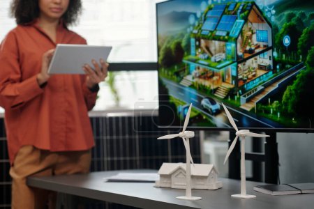 Molinos de viento de plástico blanco de pie en el escritorio contra el modelo de la casa y la pantalla con plantilla visual del nuevo proyecto empresarial durante la presentación