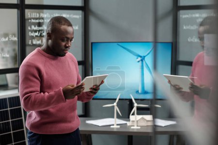 Téléchargez les photos : Jeune travailleur de bureau masculin sérieux regardant à travers les données en ligne sur l'écran de la tablette tout en se tenant contre le moulin à vent sur panneau interactif - en image libre de droit