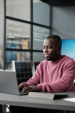 Téléchargez les photos : Travailleur de bureau afro-américain en tenue décontractée tapant sur le clavier de l'ordinateur portable et regardant l'écran tout en réseautant par lieu de travail - en image libre de droit