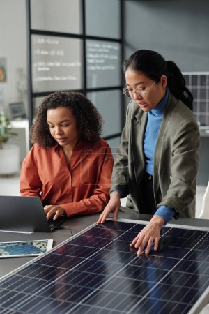 Téléchargez les photos : Jeune femme afro-américaine tapant sur un clavier d'ordinateur portable tout en étant assis à côté d'un collègue asiatique faisant la présentation du modèle de panneau solaire - en image libre de droit