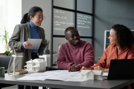 Téléchargez les photos : Trois jeunes architectes interculturels heureux discutent du nouveau projet tandis que l'un d'eux pointe vers un plan déroulé avec croquis de la maison - en image libre de droit
