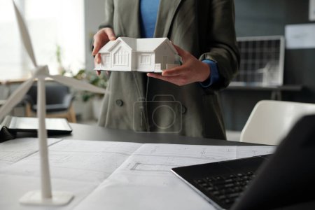 Model of house held by young unrecognizable female architect in formalwear standing by workplace with blueprints and laptop
