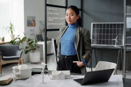 Foto de Joven empresaria asiática confiada en ropa formal mirando a la cámara durante el trabajo sobre un nuevo proyecto arquitectónico en una oficina de espacio abierto - Imagen libre de derechos