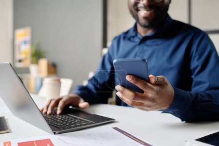 Téléchargez les photos : Crop shot de jeune gestionnaire afro-américain tenant smartphone et dactylographier sur ordinateur portable au travail dans un bureau moderne - en image libre de droit