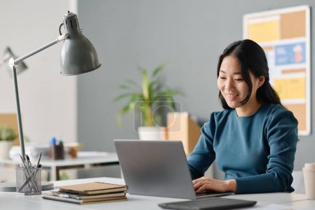 Téléchargez les photos : Joyeux jeune femme asiatique tapant sur ordinateur portable sur le lieu de travail dans un bureau moderne, espace de copie - en image libre de droit