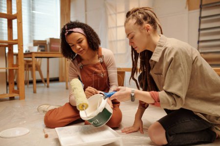 Foto de Mujer joven con la pintora sentada en el suelo junto a una colega afroamericana en ropa de trabajo que vierte pintura blanca en una bandeja de plástico - Imagen libre de derechos