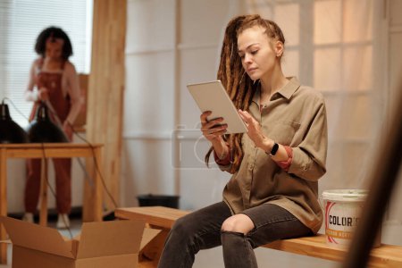 Téléchargez les photos : Jeune femme d'affaires moderne avec tablette assise sur un banc et regardant à travers des modèles en ligne avec des informations sur la rénovation - en image libre de droit