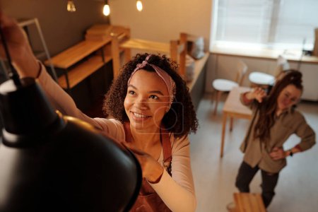 Téléchargez les photos : Jeune afro-américaine souriante propriétaire d'une nouvelle lampe suspendue au-dessus de la table pendant les travaux de réaménagement et de rénovation tout en collaborant avec elle - en image libre de droit