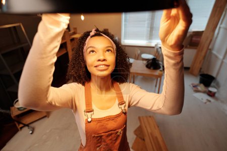Foto de Joven mujer de negocios afroamericana en ropa de trabajo colgando una nueva lámpara en el techo de la cafetería mientras hace trabajos de renovación antes de abrir - Imagen libre de derechos