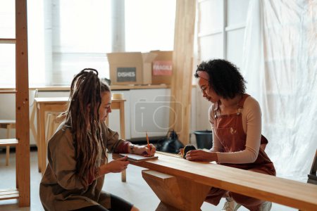 Photo for Young businesswoman with dreadlocks making notes in copybook while her colleague measuring length and width of wooden bench - Royalty Free Image