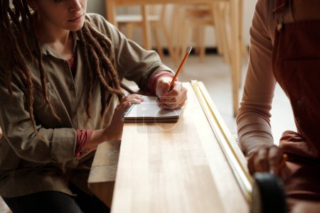 Téléchargez les photos : Jeune femme en chemise grise tenant un crayon sur la page blanche du copybook assis sur un banc en bois devant une collègue - en image libre de droit