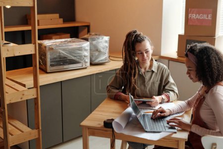 Foto de Joven mujer afroamericana en ropa de trabajo presionando las teclas del teclado del ordenador portátil mientras está sentado por la mesa junto a su colega utilizando la tableta - Imagen libre de derechos
