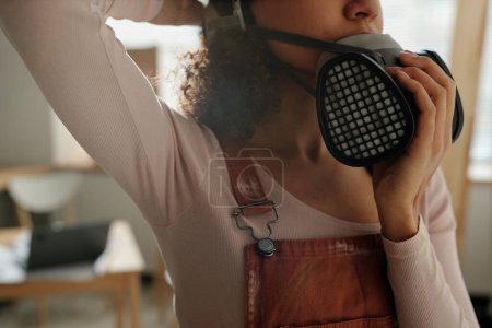 Photo for Cropped shot of young female worker in brown overalls and beige pullover putting on protective respirator before doing renovation work - Royalty Free Image