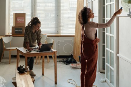 Joven mujer afroamericana en la pared de pintura de ropa de trabajo con painroller mientras está de pie contra el compañero de trabajo inclinándose sobre la mesa con el ordenador portátil