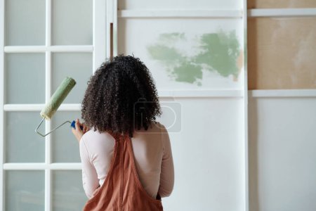 Téléchargez les photos : Vue arrière de la jeune femme afro-américaine propriétaire d'un café avec peintre à la main debout devant le mur tout en le peignant en couleur verte - en image libre de droit