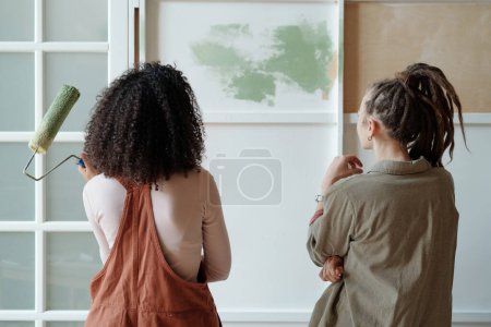 Foto de Vista trasera de dos jóvenes pintoras o diseñadoras interculturales mirando la pared de la cafetería con renovación inacabada y eligiendo el color - Imagen libre de derechos