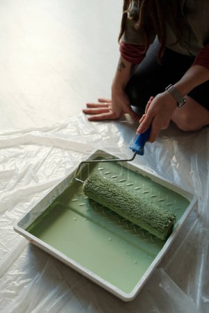 Photo for High angle of hand of young female worker sitting on knees on the floor covered by cellophane and putting paintroller in tray with green paint - Royalty Free Image