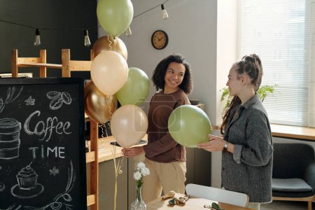 Photo for Happy young African American woman with bunch of balloons looking at colleague with dreadlocks while both decorating cafe after refit - Royalty Free Image