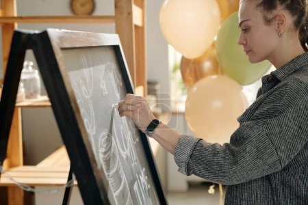 Photo for Side view of young businesswoman in grey shirt drawing advert with chalk on blackboard while preparing cafe for opening after renovation - Royalty Free Image