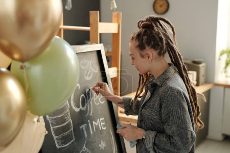 Young businesswoman with dreadlocks standing in front of blackboard and drawing advert of new cafe while preparing for opening after refit