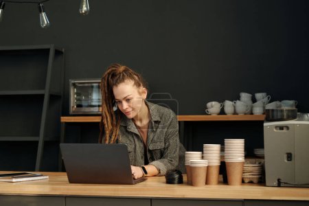 Photo for Young businesswoman in casualwear looking at laptop screen while sitting by table or counter with disposable cups and networking - Royalty Free Image