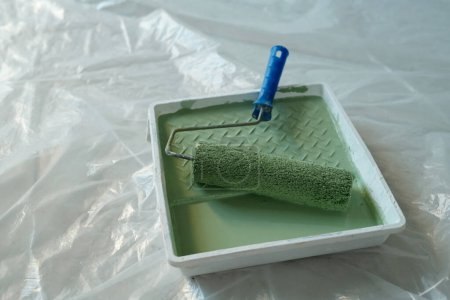 Photo for Paintroller with blue plastic handle and green paint in square tray standing on the floor covered with cellophane during renovation work - Royalty Free Image
