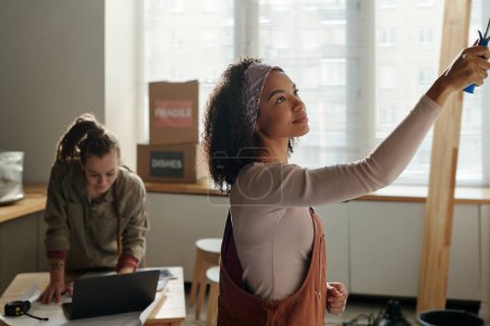 Jeune peintre féminine en salopette et pull à manches longues peignant mur de café contre son collègue regardant écran d'ordinateur portable pendant le réseau