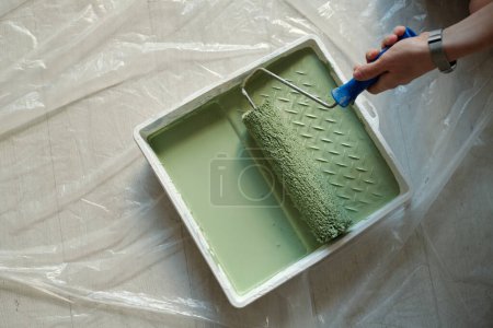 Photo for Top view of white plastic tray with green paint standing on the floor covered with cellophane and hand of woman holding paintroller - Royalty Free Image