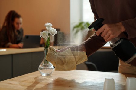 Téléchargez les photos : Main de jeune femme méconnaissable tenant une bouteille de désinfectant et le pulvérisant sur le plumeau avant de nettoyer la table après la rénovation du café - en image libre de droit