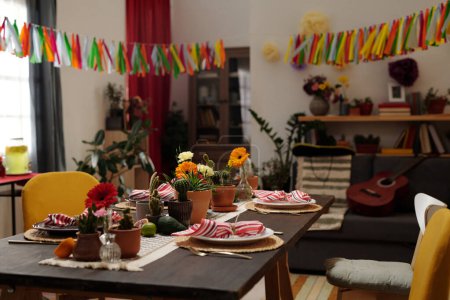 Téléchargez les photos : Partie du salon spacieux avec table de fête servie avec des fruits frais, des ustensiles de cuisine et des fleurs domestiques dans des pots de fleurs debout au centre - en image libre de droit