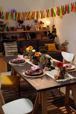 Téléchargez les photos : Table servie avec des fruits exotiques, assiettes avec des serviettes et des fleurs domestiques dans des pots de fleurs debout devant la caméra dans le salon spacieux - en image libre de droit