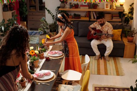 Téléchargez les photos : Femmes interculturelles dans les vêtements nationaux décoration de table avec des fleurs domestiques tandis que l'homme en casual jouer de la guitare acoustique sur le canapé - en image libre de droit