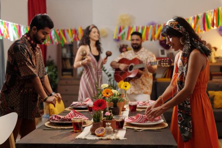 Foto de Pareja joven en ropa nacional decorando mesa de cena festiva contra sus amigos tocando la guitarra acústica y maracas en la fiesta en casa - Imagen libre de derechos