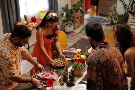 Téléchargez les photos : Groupe de jeunes amis interculturels se penchant sur la table de fête tout en la servant avec de la nourriture maison et des décorations pour la fête à la maison - en image libre de droit