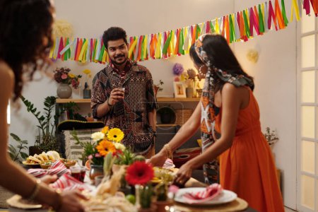 Foto de Joven hispano con vaso de tequila de pie junto a la mesa y mirando a su novia poniendo golosinas caseras junto a flores domésticas - Imagen libre de derechos