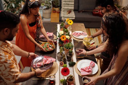 Foto de Dos parejas jóvenes en traje nacional poniendo bebidas, comida casera y platos con servilletas en la mesa festiva mientras lo sirven para la cena - Imagen libre de derechos