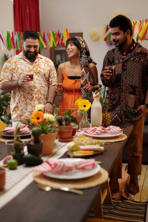 Téléchargez les photos : Trois amis joyeux en costume national trinquant avec des verres de tequila tout en se tenant à table avec des fleurs domestiques et de la nourriture maison - en image libre de droit