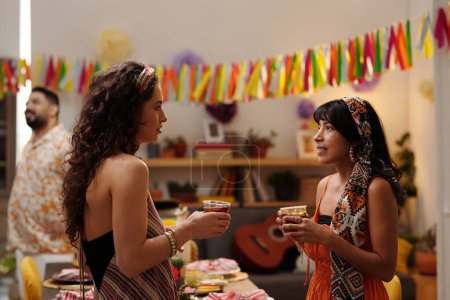 Foto de Dos chicas interculturales con bebidas en las manos mirándose durante la conversación mientras están de pie junto a la mesa de fiesta servida en la fiesta en casa - Imagen libre de derechos