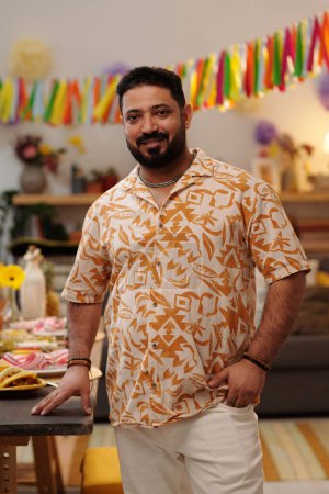Téléchargez les photos : Jeune homme souriant barbu en tenue nationale debout près de la table de fête servie avec de la nourriture maison à la fête et en regardant la caméra - en image libre de droit
