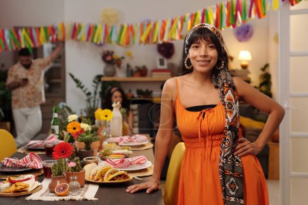 Foto de Feliz joven hispana vestida de gala nacional parada junto a la mesa festiva servida con bocadillos caseros y mirando a la cámara con sonrisa - Imagen libre de derechos