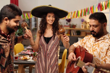 Photo for Group of happy young friends in traditional ethnic attire having fun at home party while girl in sombrero shaking maracas between guys - Royalty Free Image