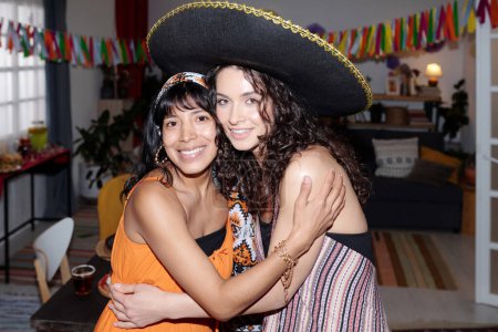 Téléchargez les photos : Les jeunes femmes interculturelles en tenue nationale debout devant la caméra dans l'étreinte tout en appréciant la fête à la maison et la célébration des vacances - en image libre de droit
