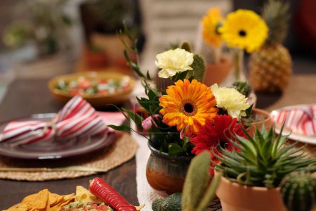 Foto de Manojo de gerberas y claveles en jarrón de pie en el centro de la mesa festiva junto a flores domésticas en macetas y aperitivos caseros - Imagen libre de derechos
