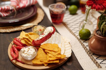 Round wooden board with nachos, red hot chili pepper and tacos standing on served festive table prepared for celebration of national holiday