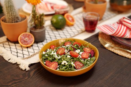 Foto de Ensalada de verduras caseras frescas en un tazón de pie sobre una mesa de madera servida con bebidas, frutas y decorada con flores domésticas - Imagen libre de derechos