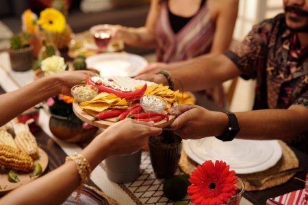 Photo for Hands of young woman passing tray with nachos, red hot chili peppers and savory sauce to male guest sitting by served festive table - Royalty Free Image