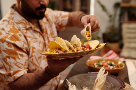 Foto de Manos de joven hispano sosteniendo plato con sabrosos tacos mexicanos y tomando uno de ellos mientras está sentado junto a la mesa festiva servida en la cena - Imagen libre de derechos