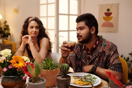 Téléchargez les photos : Jeune homme hispanique barbu avec un verre de tequila assis à côté de sa petite amie et prononçant un toast devant des amis - en image libre de droit