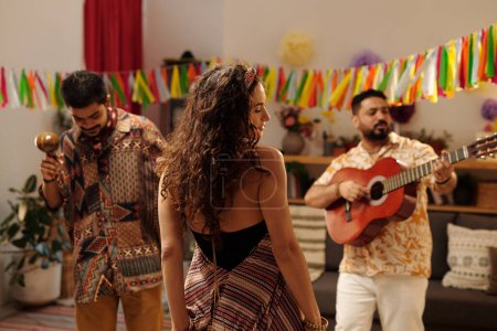 Foto de Vista trasera de una joven mujer dinámica con cabello largo oscuro y ondulado bailando frente a dos hombres hispanos tocando instrumentos musicales en la fiesta en casa - Imagen libre de derechos